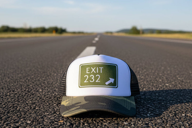 A camouflage trucker hat with a white front patch reading 'EXIT 232' and an arrow, photographed outdoors on a corrugated metal surface with greenery and flowers in the background.